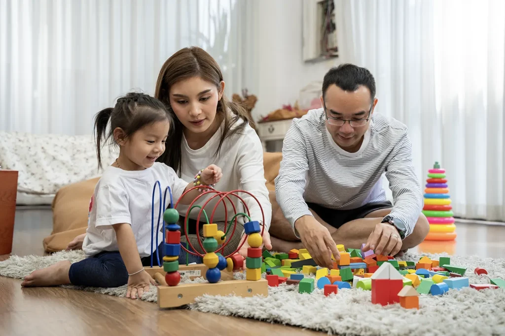 Happy young parents and their little daughter playing with wooden blocks while sitting on the living room floor; a family scene depicting parenthood and people interacting with developmental toys