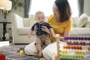 A small child happily plays with toys on the living room floor with Mom